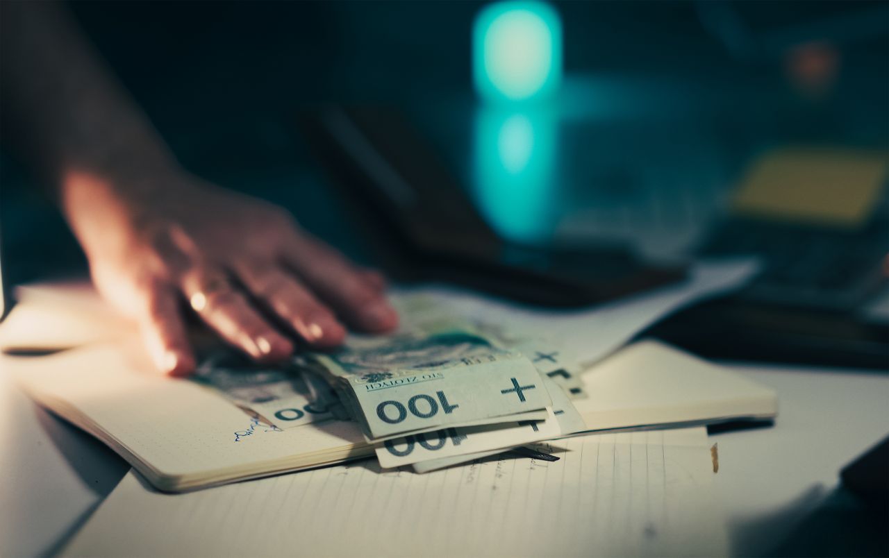 Primer plano de una mano que busca billetes en un escritorio de oficina con un cuaderno y una calculadora.
